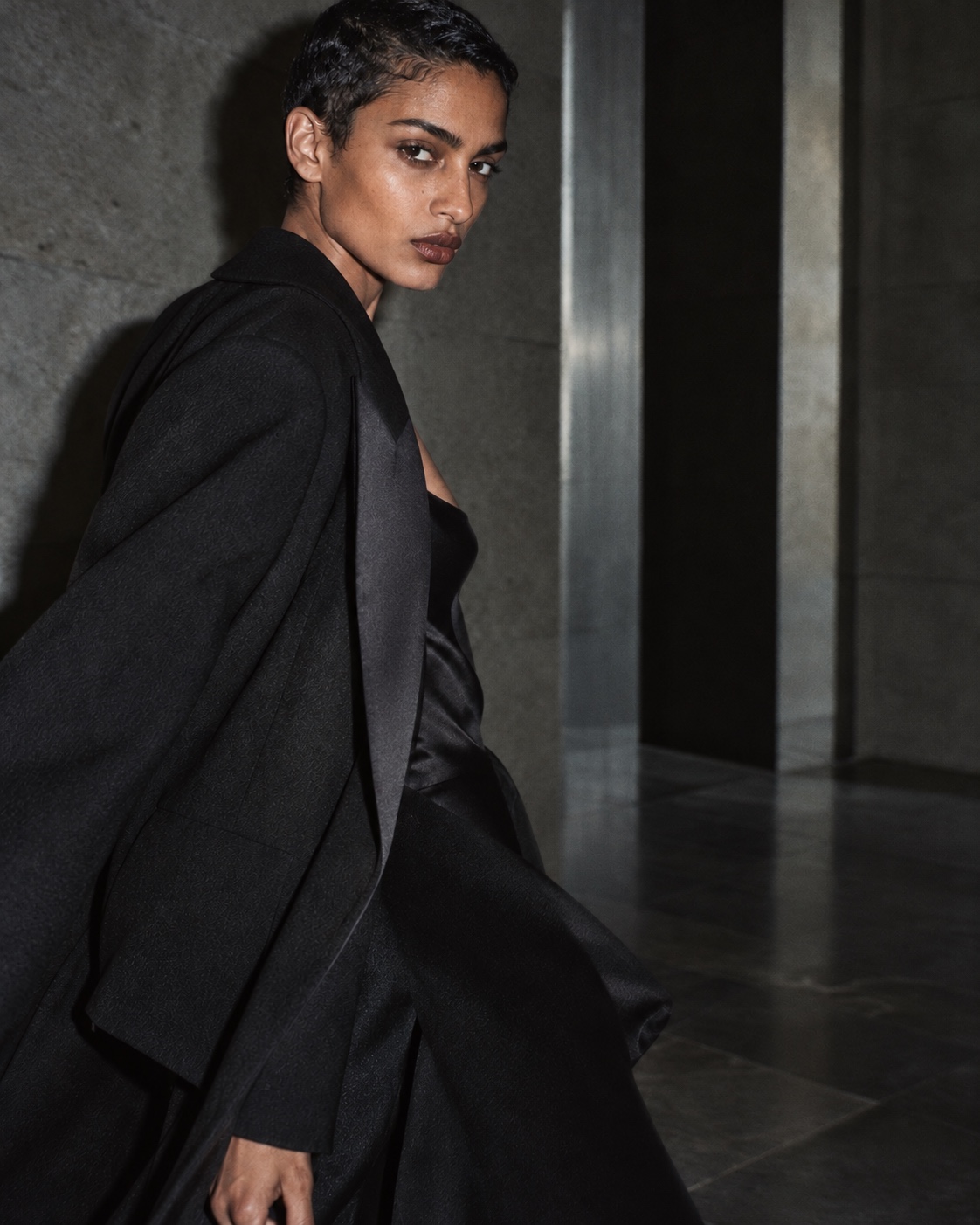 A woman in a sculptural black coat turns toward the camera in a gray stone interior.